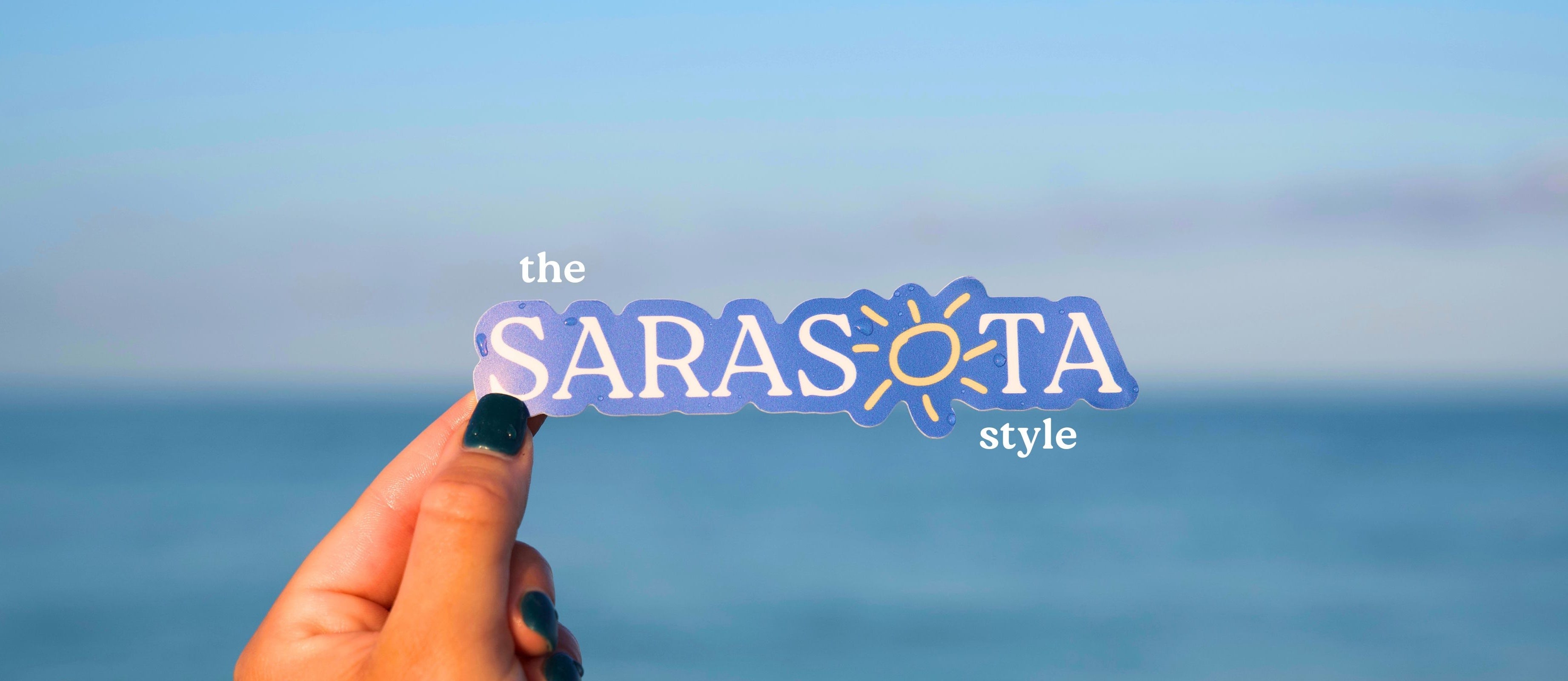 Hand holding a 'the SARASOTA style' sicker with a blue sky and ocean