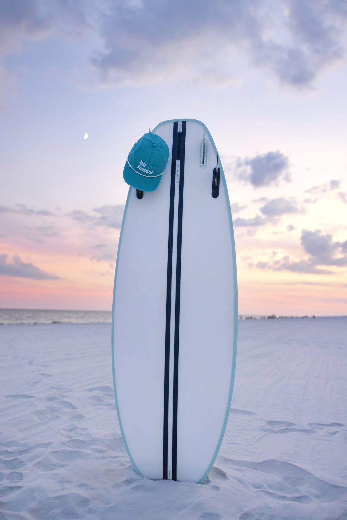 Beach Happy cap on a surfboard in the sand at sunset