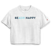 A light heather gray short-sleeve crop top t-shirt featuring the text 'BEACH HAPPY' printed across the chest, with 'BEACH' in dark blue and 'HAPPY' in light blue. A small red logo is visible on the right sleeve.